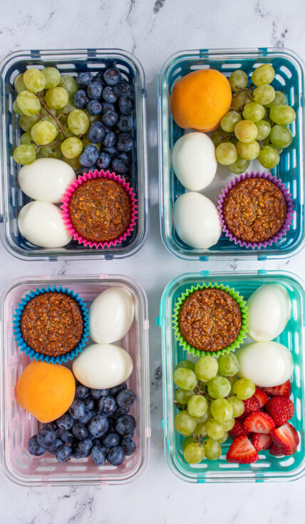 Four breakfast boxes on a tabletop each with hard boiled eggs, muffin, and fruit.
