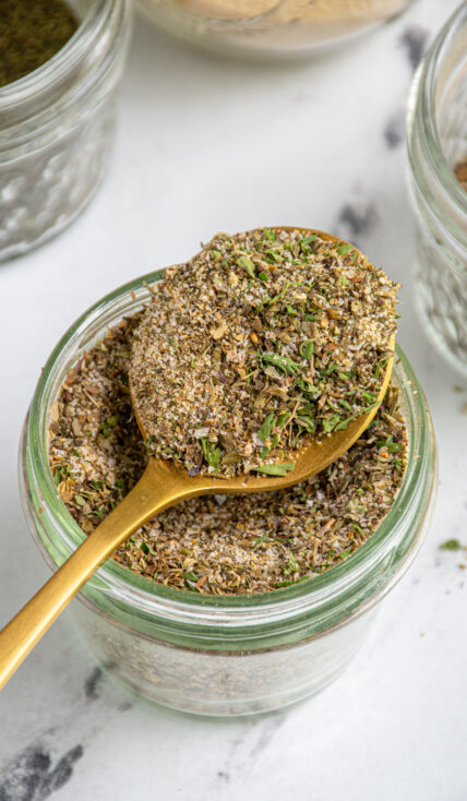 A jar with a spoon resting on top filled with homemade greek seasoning recipe.