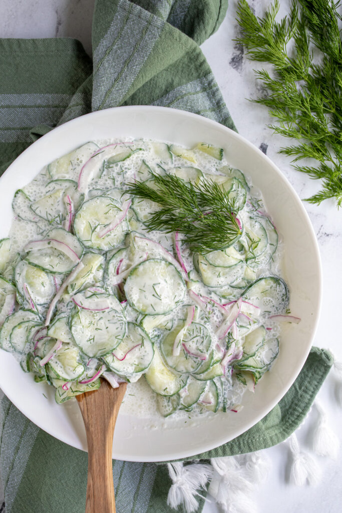 A white serving bowl with creamy cucumber salad and fresh dill garnish next to a green kitchen towel.