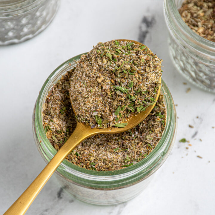 A glass jar with a spoon resting on top with the spoon full of homemade greek seasoning.