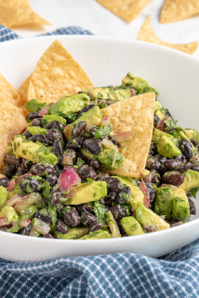 A white bowl with avocado black bean salsa and a tortilla chip full of black beans and avocado chunks.