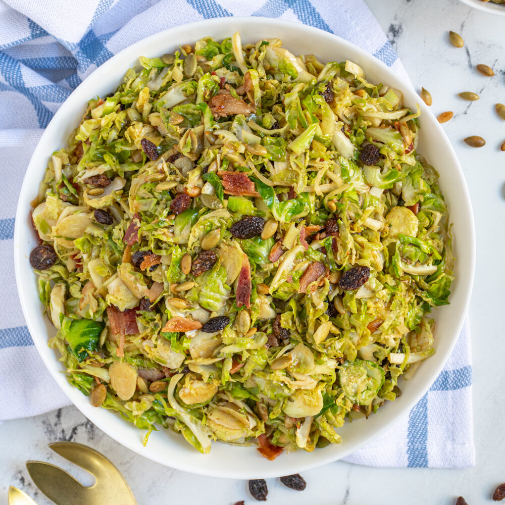 Overhead view of a large white bowl full of warm balsamic brussel sprout salad with pumpkin seeds, raisins, and bacon.