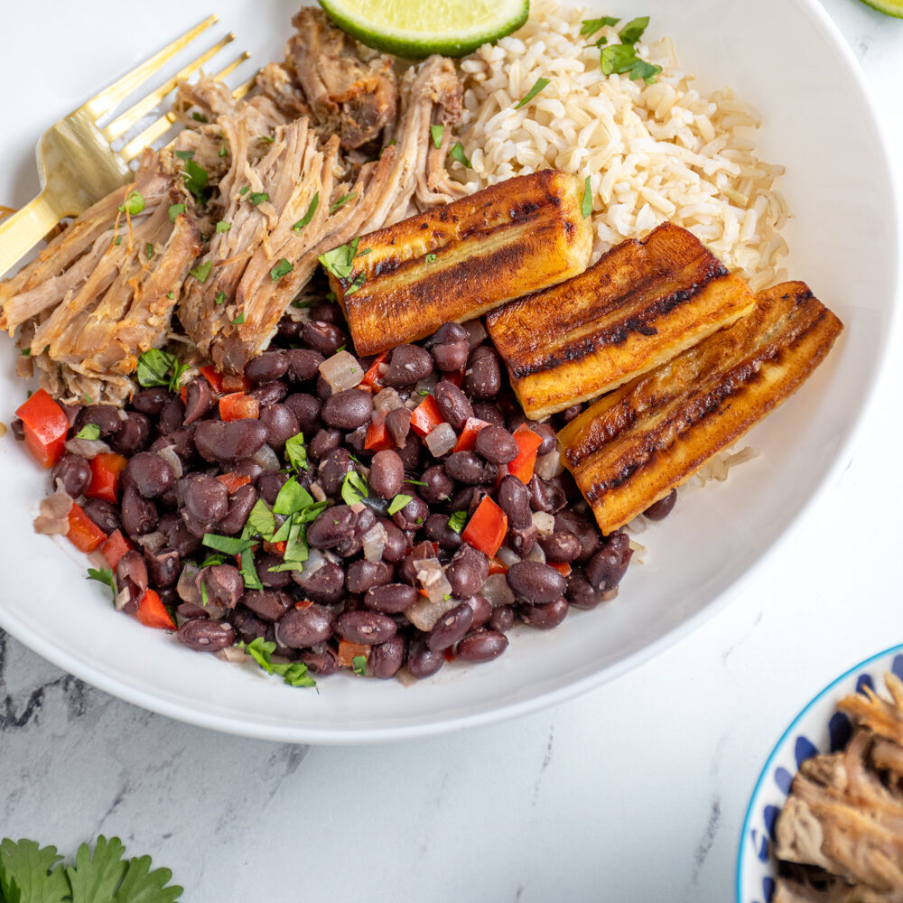 A cuban bowl filled with rice, mojo pork, cuban black beans, and fried bananas garnished with cilantro and lime wedges.