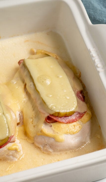 Close up of a baked cubano chicken in a casserole dish with deli meats, pickles, and swiss cheese.