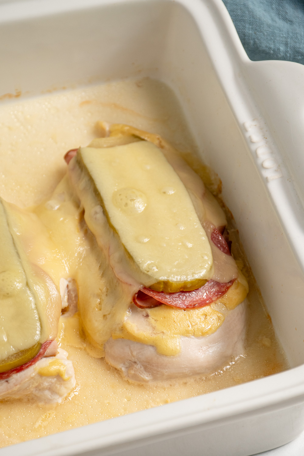 Close up of a baked cubano chicken in a casserole dish with deli meats, pickles, and swiss cheese.