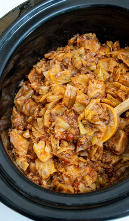 A black crockpot with unstuffed cabbage rolls being scooped with a wooden spoon.