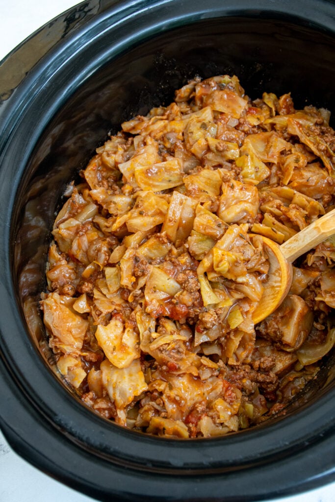 A black crockpot with unstuffed cabbage rolls being scooped with a wooden spoon.