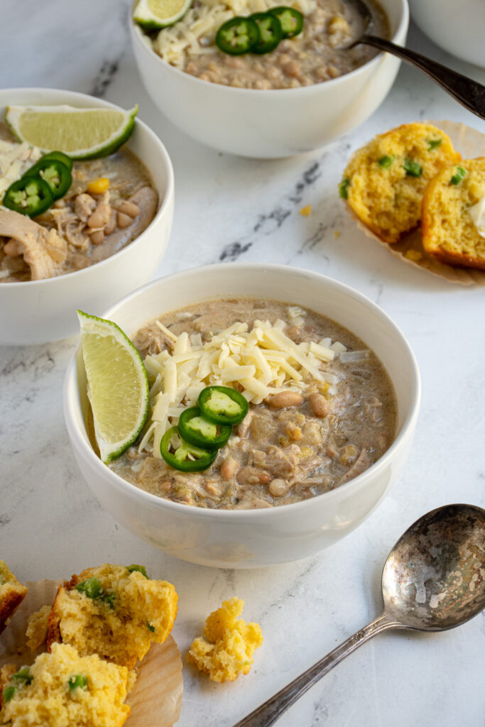 A white bowl next to a cornbread muffin filled with white chicken chili and garnished with sharp cheddar, jalapeños, and a lime wedge.