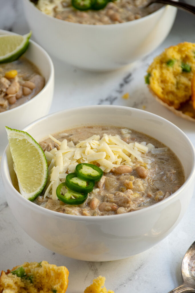 Close up of a white bowl filled with crockpot white chicken chili garnished with sharp cheddar, jalapeños, and limes wedges.