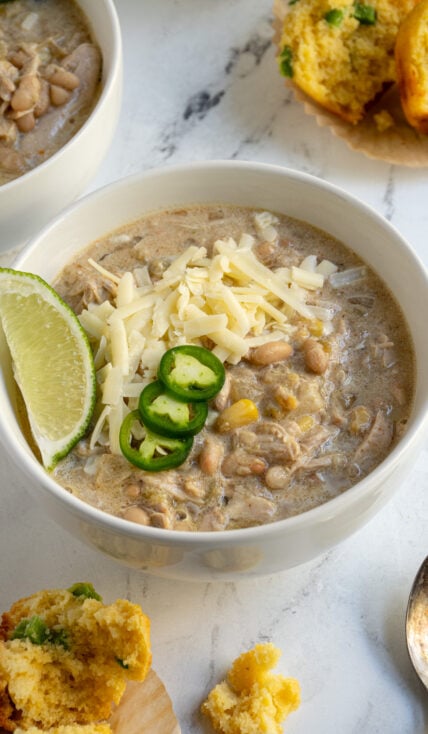 A white bowl filled with crockpot white chicken chili garnished with sharp cheddar, jalapeños, and lime wedge.