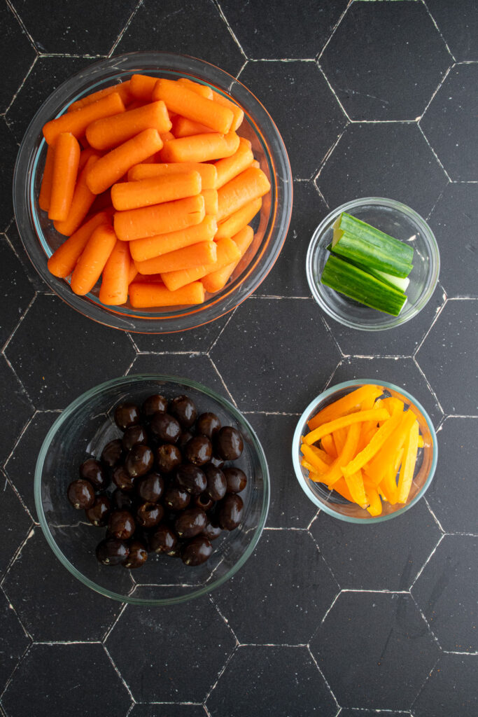 Ingredients for a pumpkin veggie tray laid out on a black surface including carrots, cucumber, orange bell pepper, and black olives.