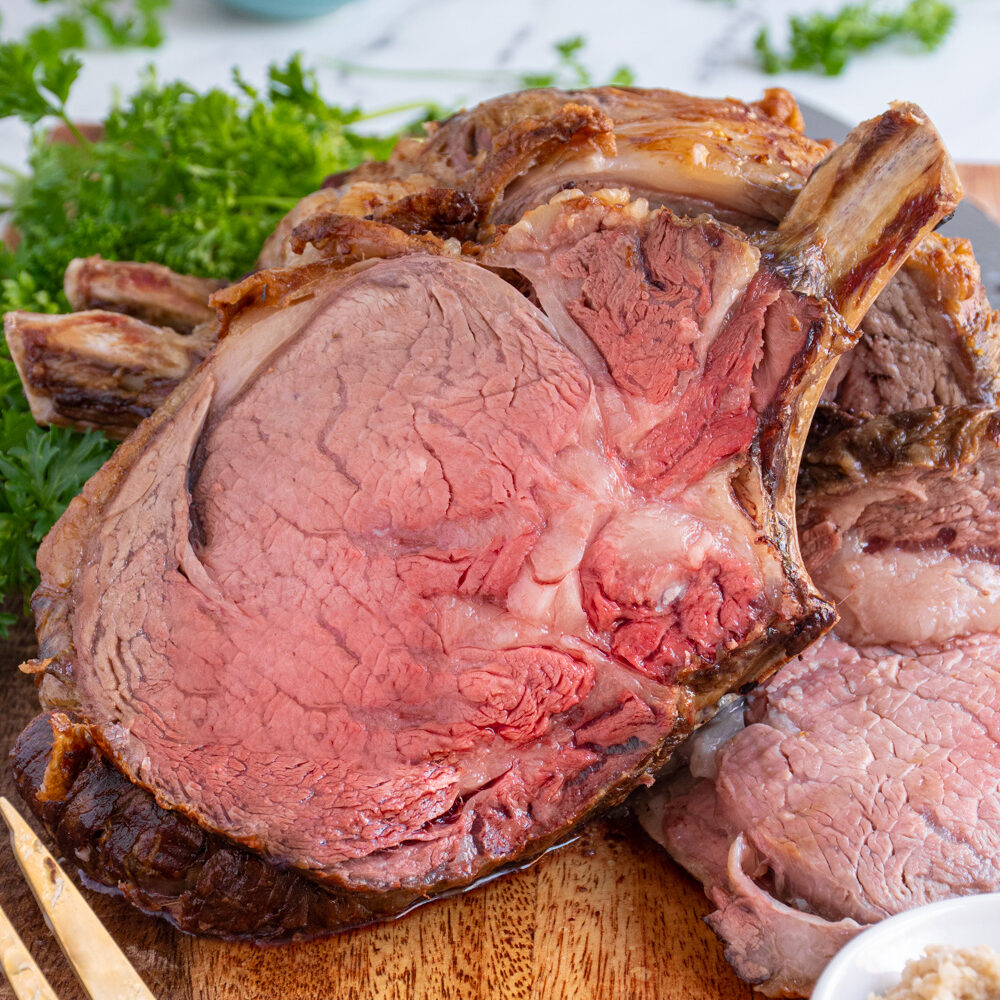 A large slice of prime rib on a cutting board leaned up against the rest of the roast showing off a red tender inside.