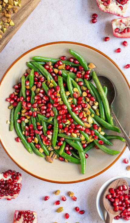 A brown serving bowl filled with bight green beans and topped with red pomegranate seeds and chopped pistachios.