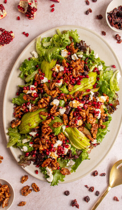 A large platter covered in a festive winter salad with candied walnuts, pomegranates, dried cranberries, pears, and goat cheese.