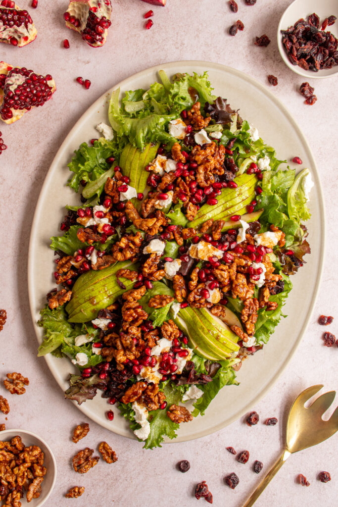 A large platter covered in a festive winter salad with candied walnuts, pomegranates, dried cranberries, pears, and goat cheese.
