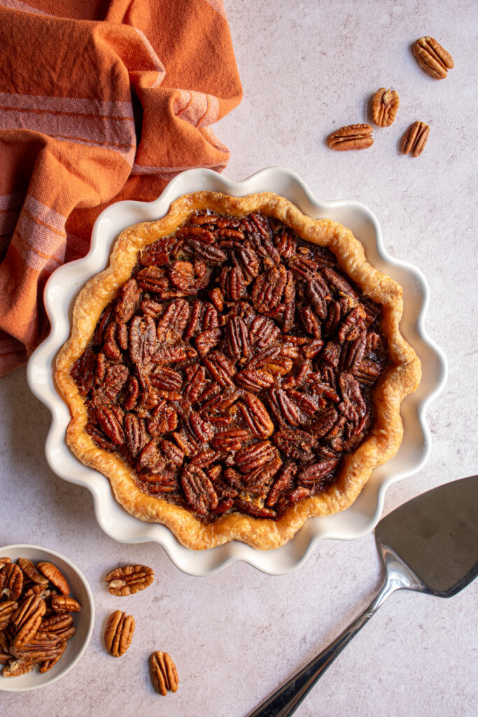Flatlay of a pie plate with a bourbon pecan pie surrounded by pecans and a pie server.
