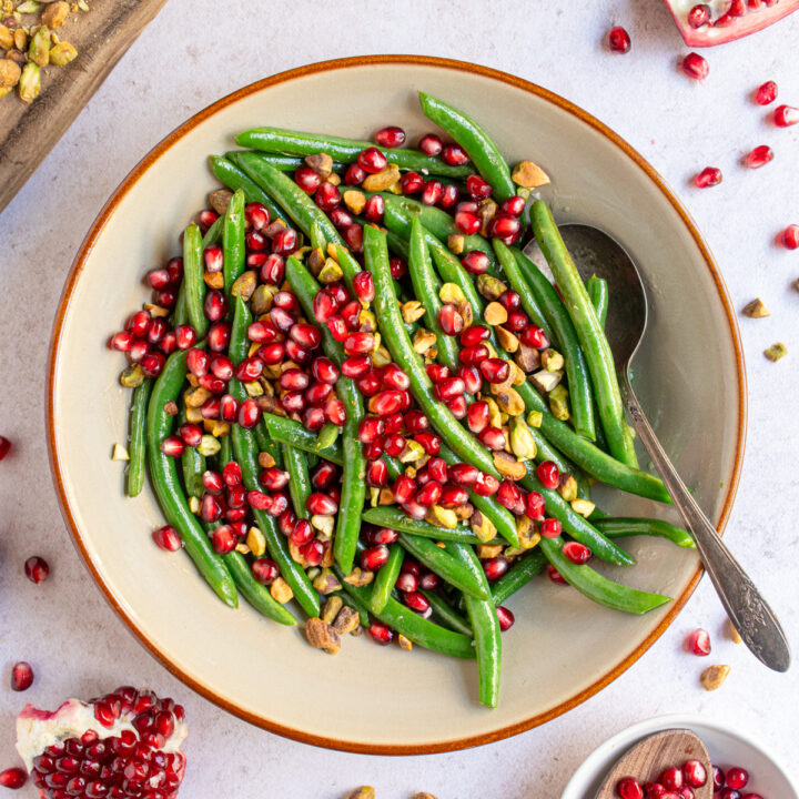 A bowl surrounded by pomegranate pieces, in the bowl in green beans topped with pomegranate seeds and chopped pistachios.