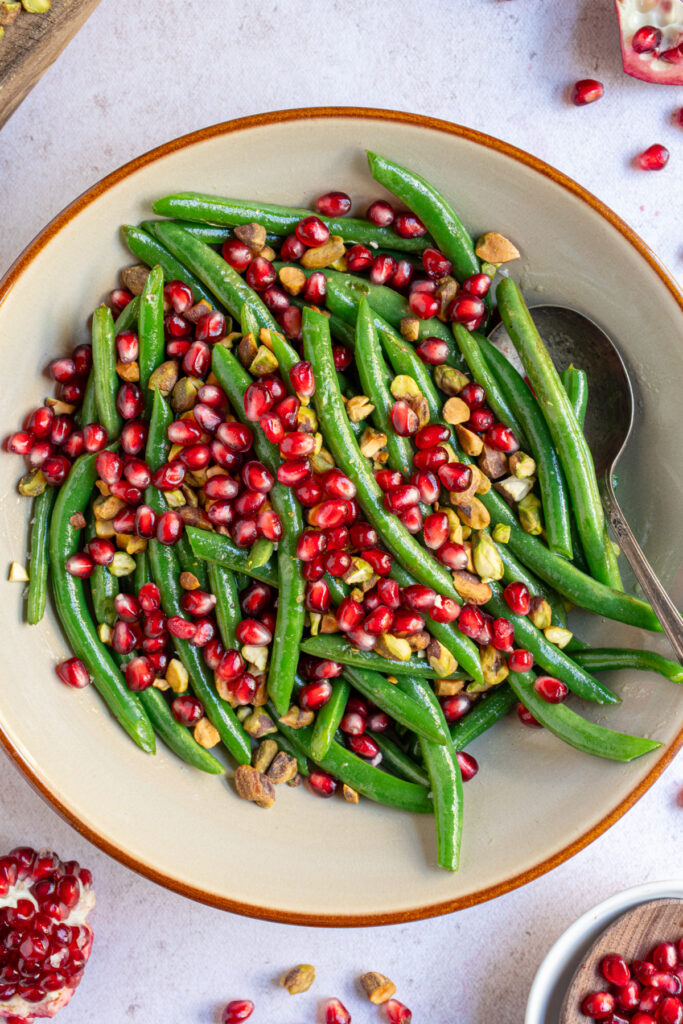 A brown bowl filled with bight sautéed green beans topped with bright red pomegranate seeds and shopped pistachios.