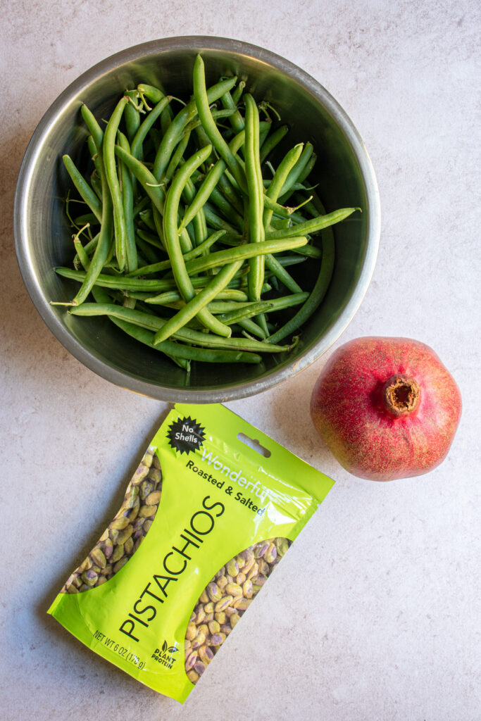 Green beans, a pomegranate, and pistachios laid out on a tabletop.