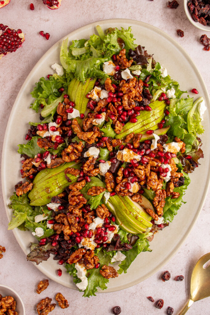 Over view of a large salad on a platter with candied walnuts, pears, pomegranate seeds, cranberries, and goat cheese.