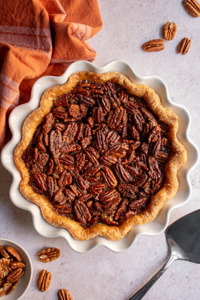 Overhead photo of a bourbon pecan pie on a white pie plate next to pecans and a pie server.