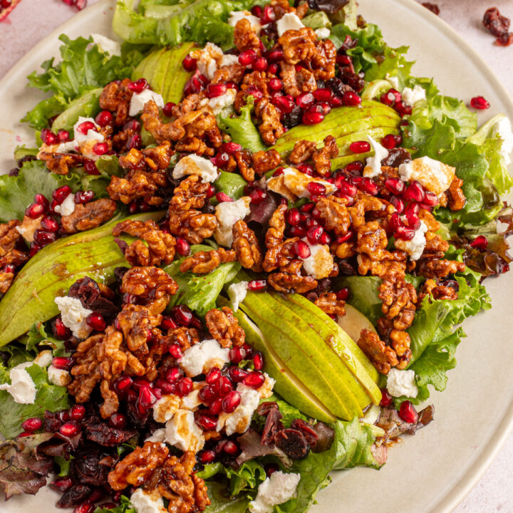 A large white platter with a winter salad topped with candied walnuts, pears, pomegranate seeds, dried cranberries, and goat cheese.
