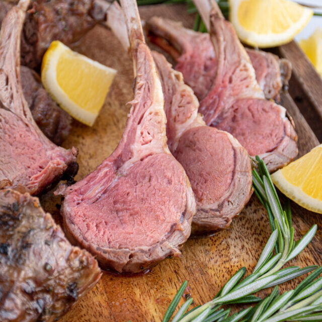 A few pieces of cut grilled rack of lamb on a wooden cutting board showing off the rosy interior color of the grilled lamb meat.