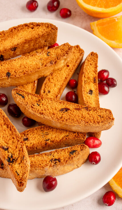 A white plate with a pile of Orange Cranberry Biscotti surrounded by cranberries and orange wedges.