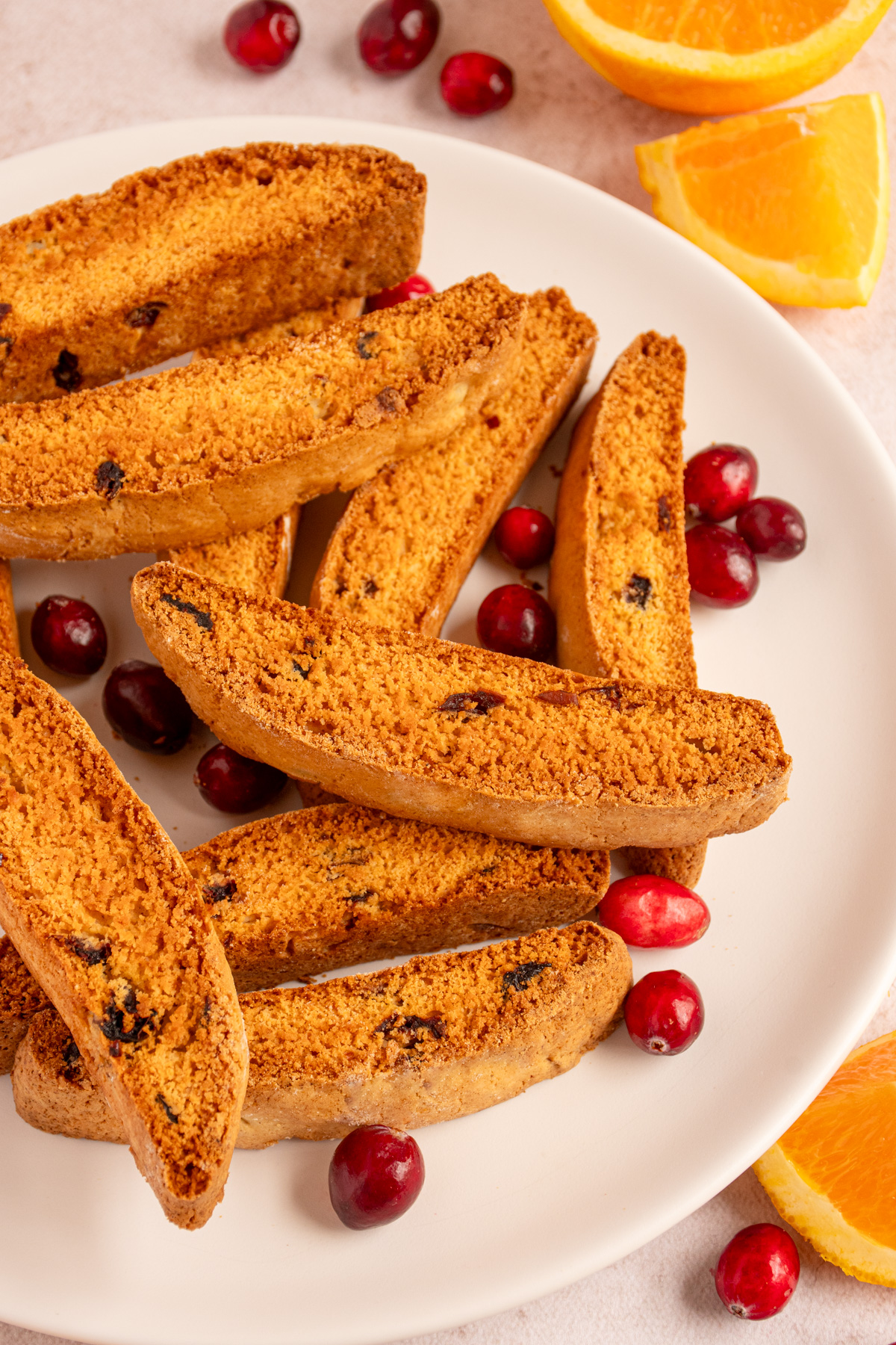 A white plate with a pile of Orange Cranberry Biscotti surrounded by cranberries and orange wedges.