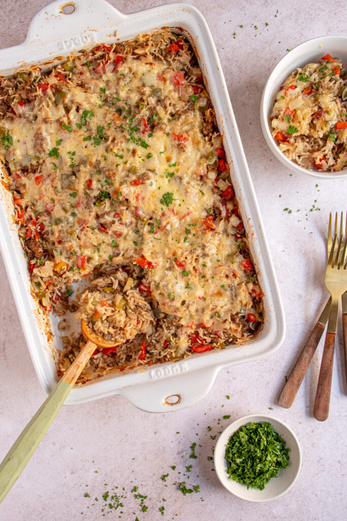 Over head view of a baking dish and bowl with stuffed pepper casserole with a wooden spoon showing the cooked rice, ground beef, and bell peppers.