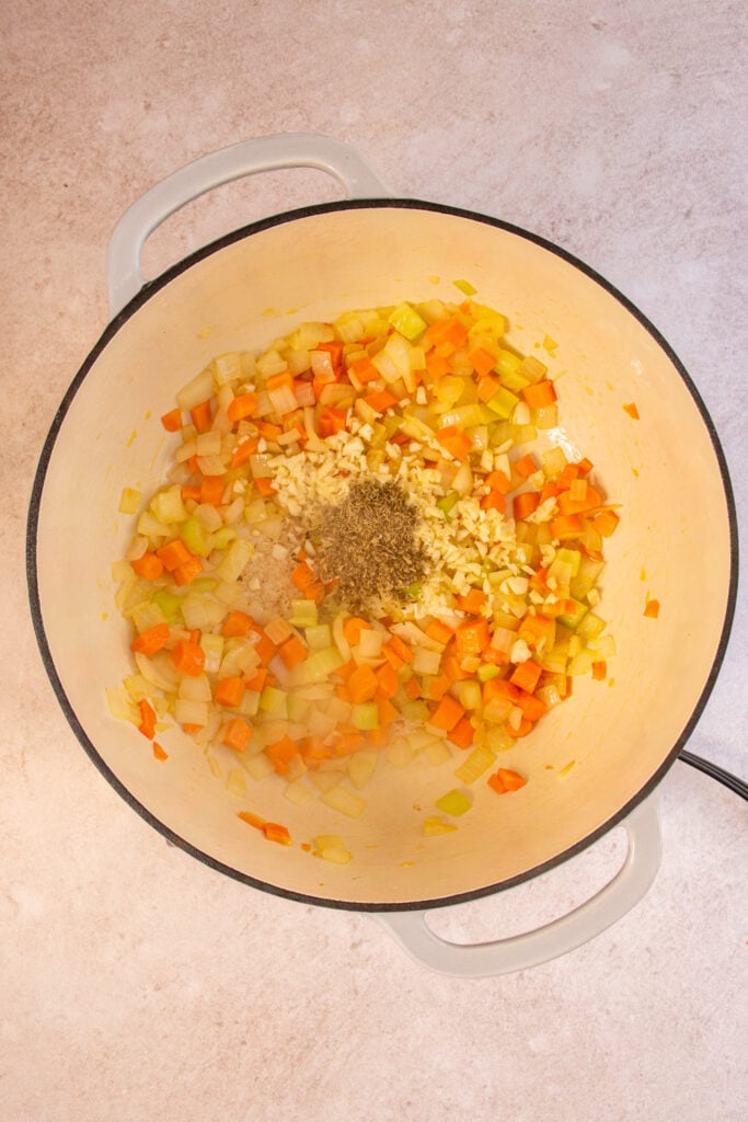 A large dutch oven with veggies inside it and dried herbs and garlic being added.