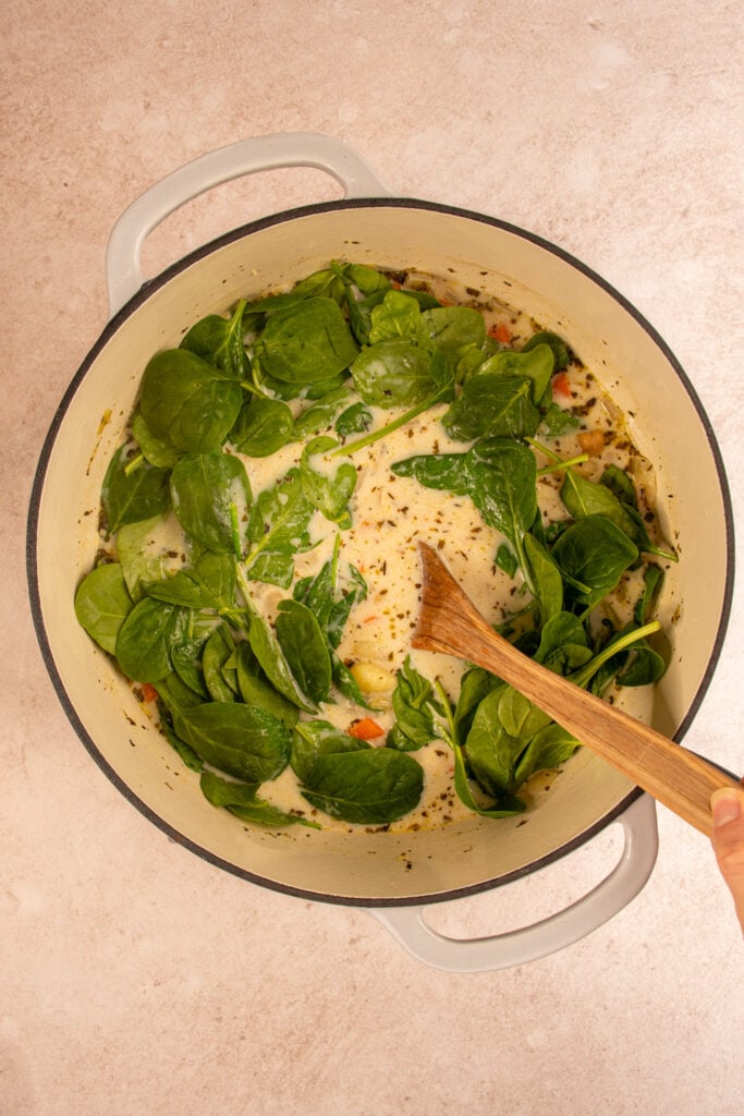 A large dutch oven where a few handfuls of baby spinach are being added to a large dutch oven.