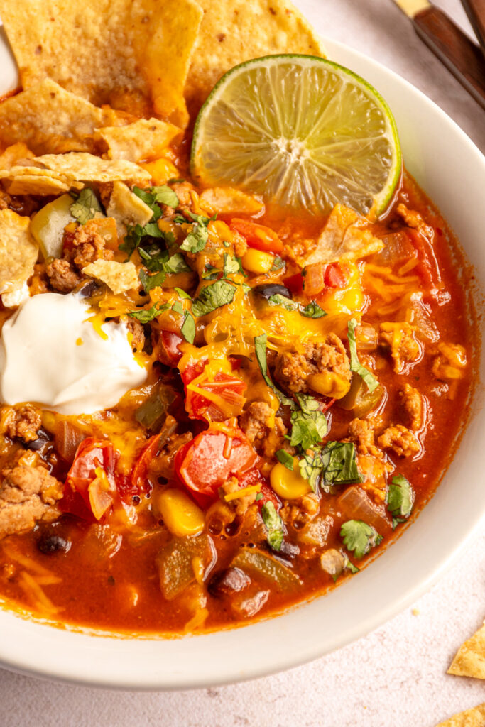Close up of a white bowl filled with turkey taco soup topped with cheddar cheese, sour cream, cilantro, tortilla chips, and lime.