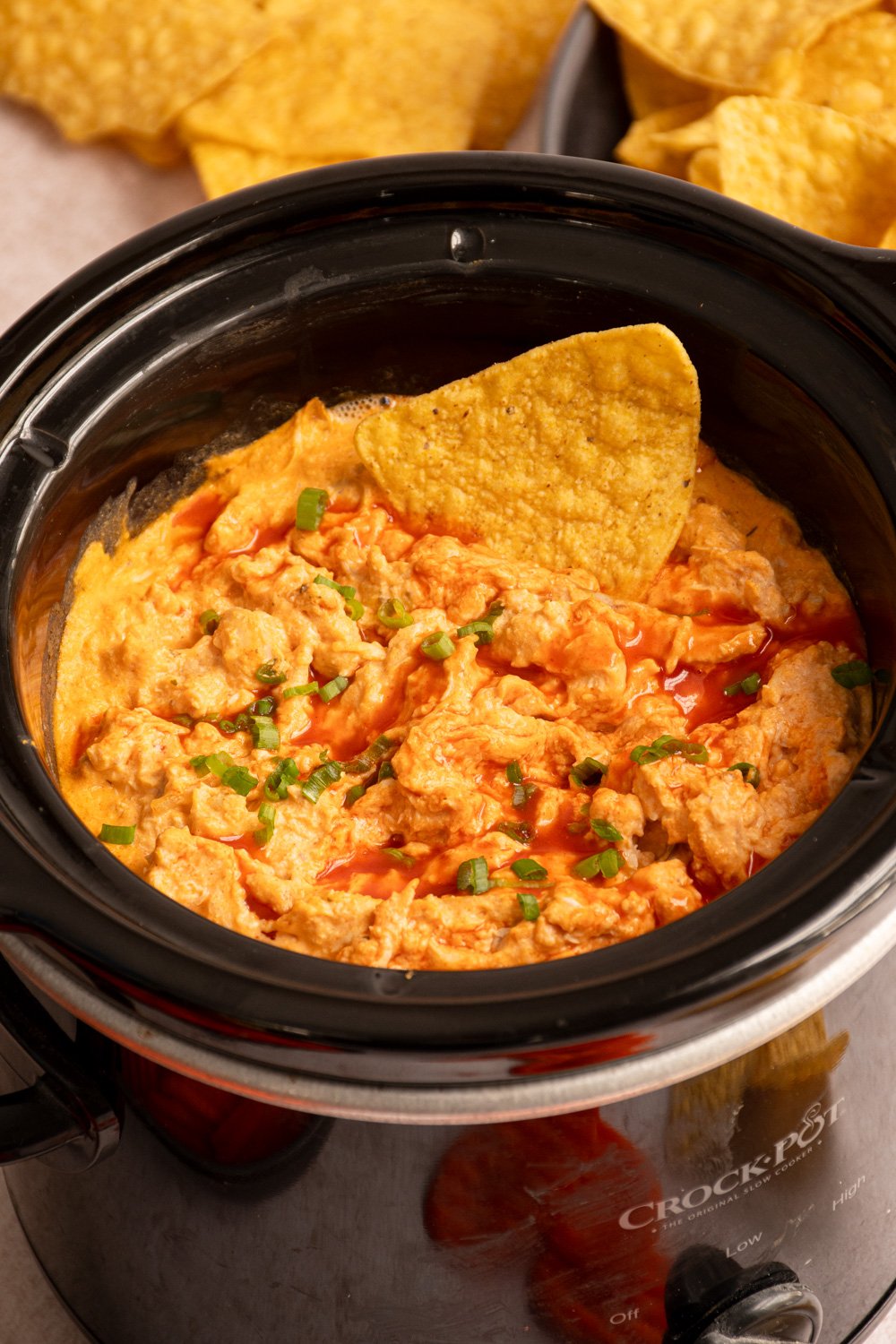 A small slow cooker filled with buffalo chicken dip garnished with green onions and a chip dipped in.