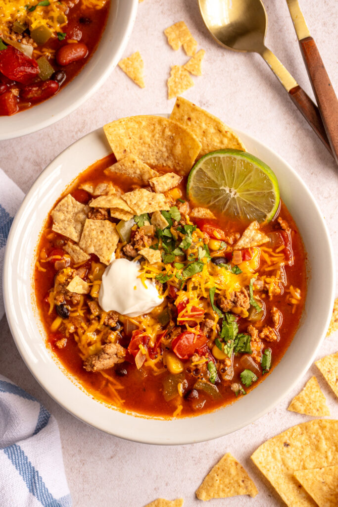 A white bowl filled with healthy ground turkey taco soup with shredded cheese, sour cream, cilantro, and tortilla chips.