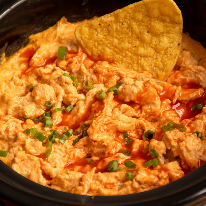 Buffalo chicken dip in a small slow cooker with a chip being dipped in.