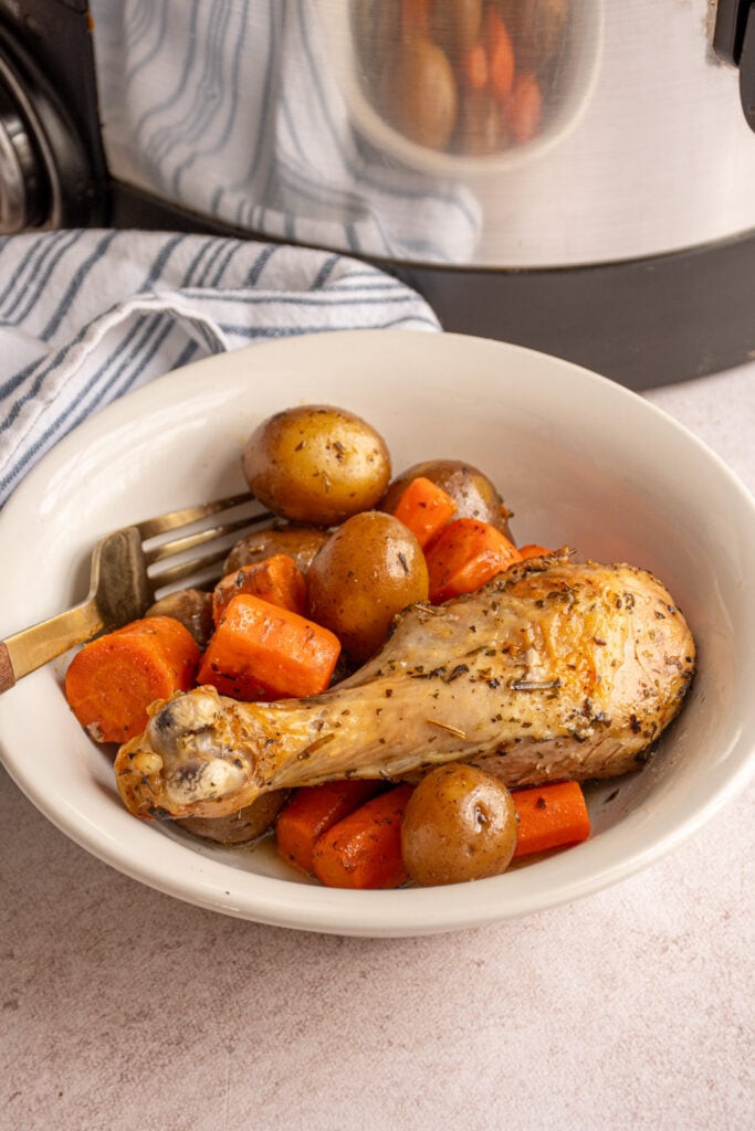 A chicken legs with carrots and baby potatoes in a white bowl with a fork.