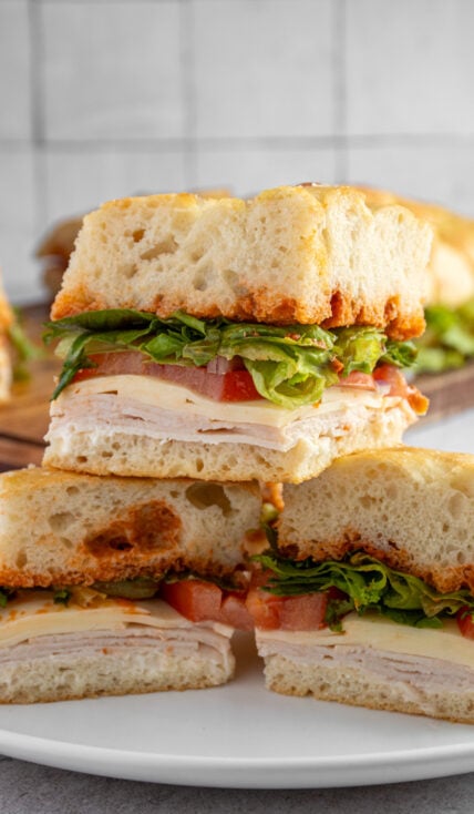 Individual pieces of focaccia turkey sandwich layered on a plate showing off layers of turkey, cheese, tomato, lettuce, and sun-dried tomato pesto in between layers of focaccia bread.