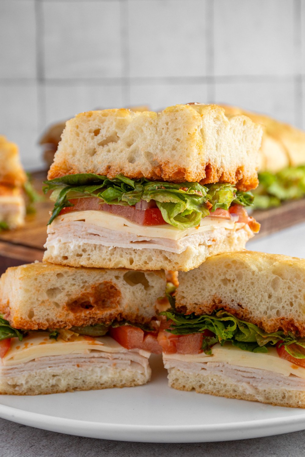 Individual pieces of focaccia turkey sandwich layered on a plate showing off layers of turkey, cheese, tomato, lettuce, and sun-dried tomato pesto in between layers of focaccia bread.