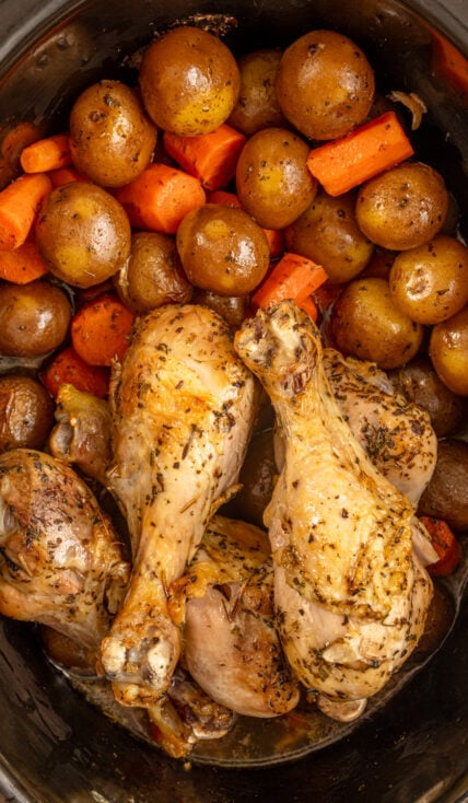 A black crockpot with cooked carrots, potatoes, and chicken legs.
