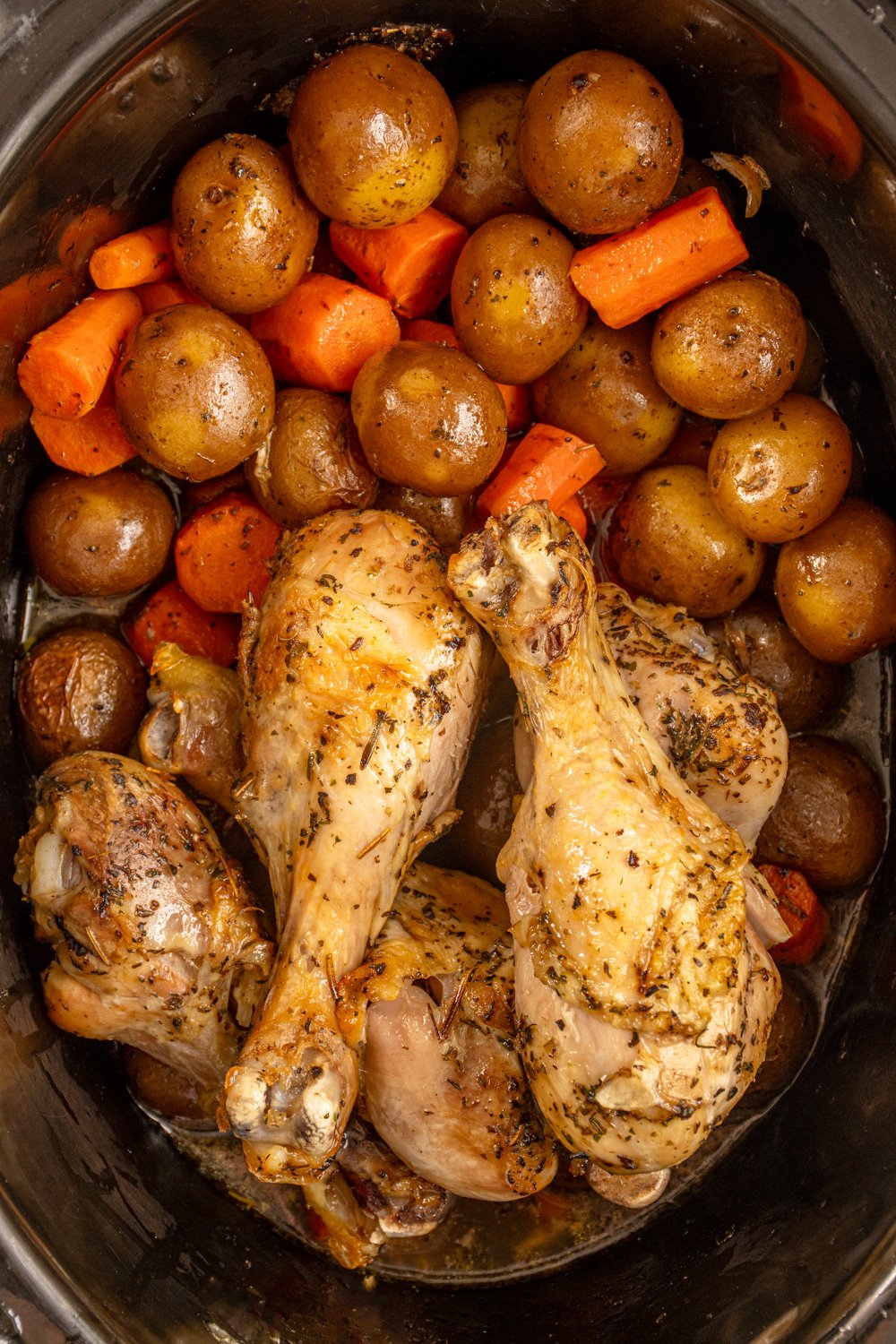 A black crockpot with cooked carrots, potatoes, and chicken legs.