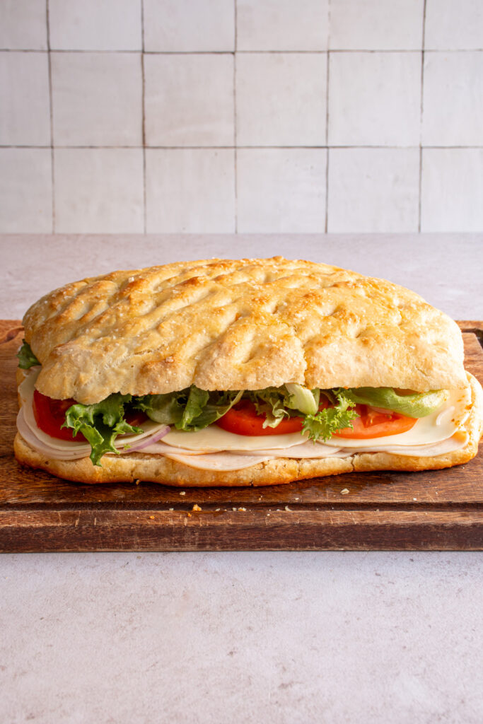 A large loaf of focaccia turned into a sandwich to feed a crowd on a wood cutting board with layers of turkey, cheese, tomato, lettuce, and sun-dried tomato pesto between layers of focaccia bread.