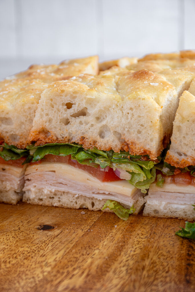 Focaccia Turkey Sandwich on a wood cutting board with layers of turkey, cheese, tomato, lettuce, and pesto between focaccia bread.