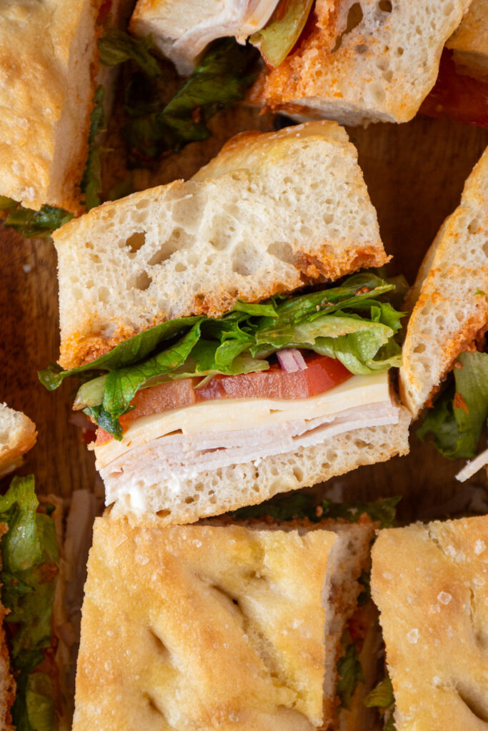 A piece of focaccia turkey sandwich showing layers of turkey, cheese, tomato, lettuce, and sun-dried tomato pesto between focaccia bread.