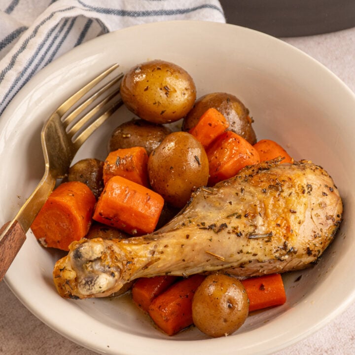 A white bowl with cooked potatoes and carrots with a chicken leg.