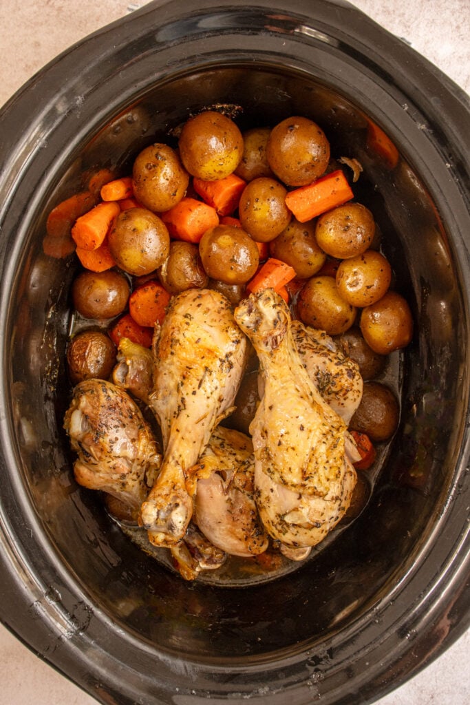 A black crockpot filled with cooke carrots and baby potatoes with chicken legs.