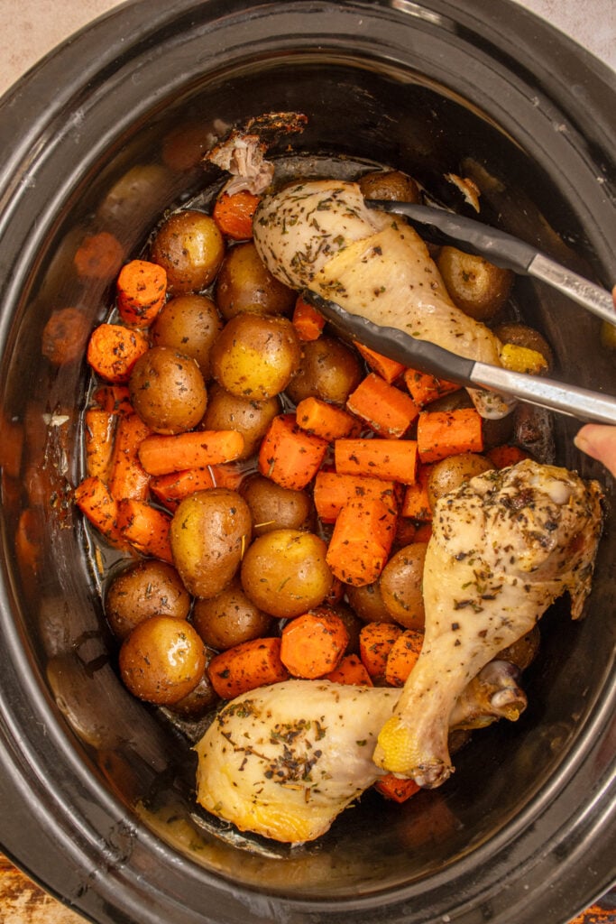 Taking a cooked chicken leg out of the crockpot showing the cooked vegetables underneath.