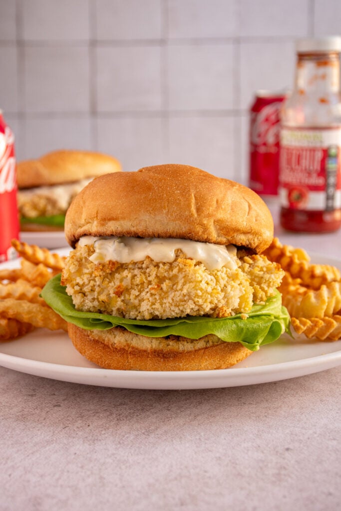 A baked fish sandwich on a plate in between soft buns with tartar sauce and lettuce.