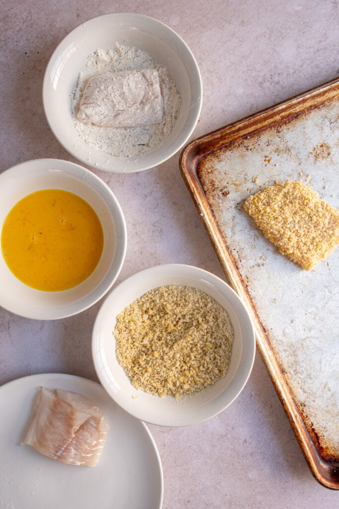 Adding a fish fillet to the flour mixture in a bowl to coat it as the first part of the breading process.