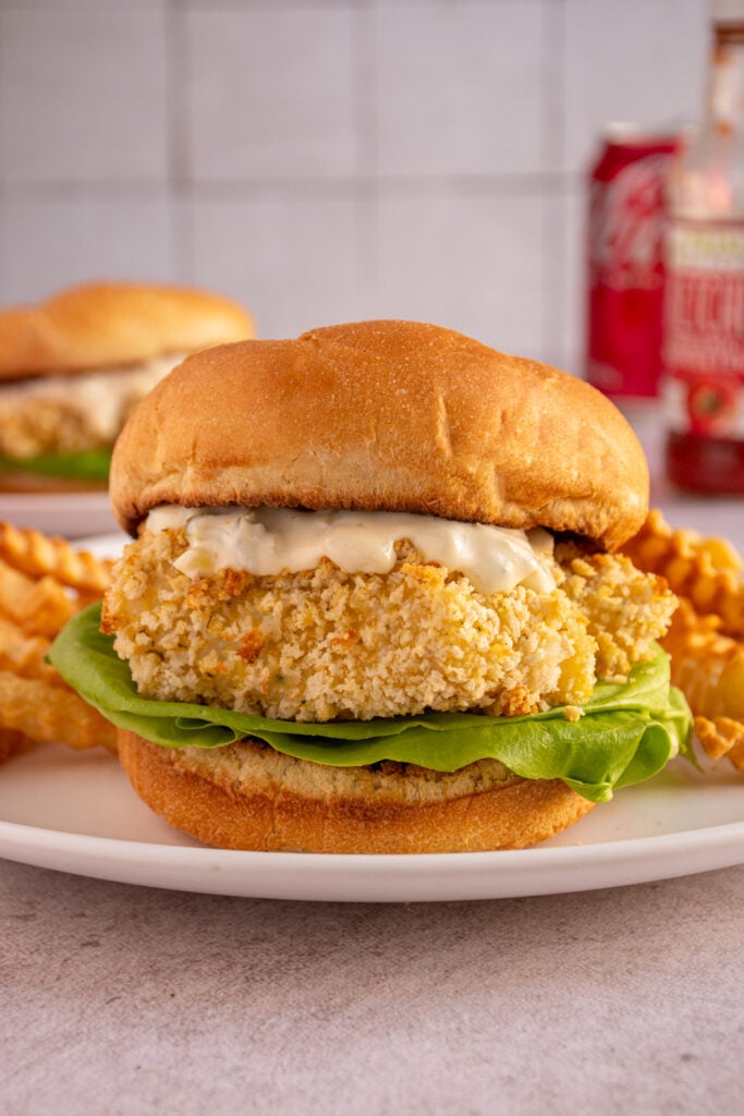 A baked fish sandwich on a white plate in between soft buns with tartar sauce and lettuce.